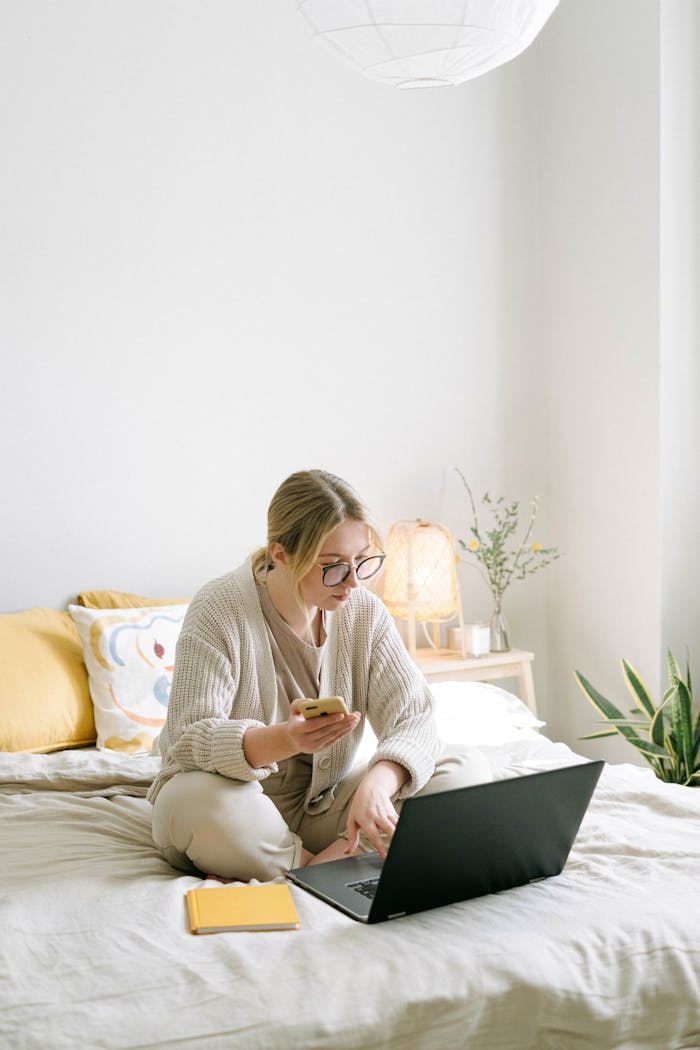 A woman working remotely from home sitting on a bed using a laptop and phone in a cozy bedroom.