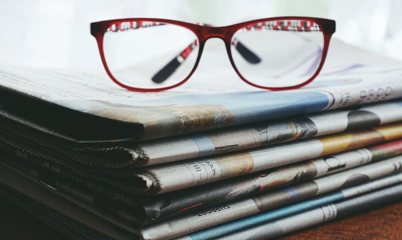 Red eyeglasses resting on a stack of newspapers symbolize reading and information.